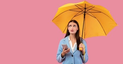 Eine junge Frau steht mit einem Regenschirm vor einer einfarbigen Wand. Sie hat ihr Handy in der Hand und blickt skeptisch zum Himmel. Ein Symbolbild für Preventive Maintenance.